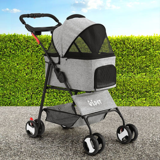 A pet stroller with a gray heathered fabric body and black mesh canopy, shown outdoors on a paved surface with a green hedge and blue sky in the background. The stroller has a black frame, black handlebar, and white accents on the wheels and a small logo on the front fabric pocket.